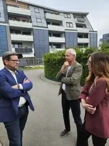 Bundestagsabgeordneter Lars Rohwer im Gespräch mit FASA‑AG‑Geschäftsführer Ulrich Hintzen und einer Kollegin vor einem modernen Mehrfamilienhaus mit solaraktiver Fassadenverkleidung.