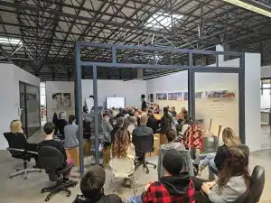Im Showroom der FASA AG nehmen zahlreiche Mitarbeitende und Auszubildende an einer Sicherheitsschulung teil: Sie sitzen dicht an dicht auf Stühlen und Hockern um einen langen Holztisch, während vorne eine Referentin bzw. ein Referent eine Präsentation zu Arbeitssicherheit auf einem großen Bildschirm erklärt; an den Wänden hängen Projektfotos und Infotafeln des Unternehmens.