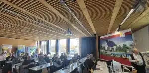 Im Seminarraum der Hochschule Weihenstephan-Triesdorf verfolgt ein vollbesetztes Plenum die Jahreshauptversammlung des Sonnenhaus-Instituts: Vorne rechts präsentiert ein Referent vor einem großen Banner „Das Sonnenhaus“ mit solarthermischem Gebäude, während die Teilnehmenden an langen Tischen mit Getränken und Unterlagen sitzen; im Hintergrund informieren mehrere Roll-ups über Projekte und Inhalte rund um erneuerbare Energien.