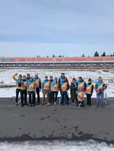 Gruppenfoto vom FASA-Sporttag in Oberwiesenthal: Eine größere Gruppe von Mitarbeitenden und Angehörigen steht auf der teilweise verschneiten Biathlonanlage, alle tragen orange-blaue Startnummernwesten, im Hintergrund sind die beschilderten Schießstände sowie Bandenwerbung der Sparkasse und weiterer Partner zu sehen.