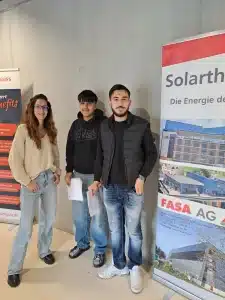 Zwei Schüler der Marianne-Brandt-Oberschule neben dem Roll-up-Banner der FASA AG auf einer Minimesse in einer Schule: Oben Firmenlogo mit Schriftzug „FASA AG“, daneben stilisierte Grafik eines Vogels, darunter der Slogan „Bau mit uns auf deine Zukunft.“ Auflistung verschiedener Ausbildungsberufe im Bau- und Technikbereich (u. a. Anlagenmechaniker SHK, Beton- und Stahlbetonbauer, Hochbaufacharbeiter, Industriekaufleute, Maurer, Zimmerer) sowie Vorteile der Ausbildung wie Vergütung, Jahresprämie, ÖPNV-Zuschuss, Teamgeist und Weiterbildungsmöglichkeiten. Rechts ist ein modernes Gebäude abgebildet sowie unten rechts eine Person beim Mauern großer Ziegelsteine, im Vordergrund liegen Infomaterialien und Stifte auf einem Tisch.