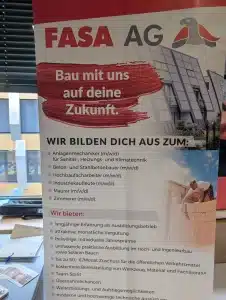 Roll-up-Banner der FASA AG auf einer Minimesse in einer Schule: Oben Firmenlogo mit Schriftzug „FASA AG“, daneben stilisierte Grafik eines Vogels, darunter der Slogan „Bau mit uns auf deine Zukunft.“ Auflistung verschiedener Ausbildungsberufe im Bau- und Technikbereich (u. a. Anlagenmechaniker SHK, Beton- und Stahlbetonbauer, Hochbaufacharbeiter, Industriekaufleute, Maurer, Zimmerer) sowie Vorteile der Ausbildung wie Vergütung, Jahresprämie, ÖPNV-Zuschuss, Teamgeist und Weiterbildungsmöglichkeiten. Rechts ist ein modernes Gebäude abgebildet sowie unten rechts eine Person beim Mauern großer Ziegelsteine, im Vordergrund liegen Infomaterialien und Stifte auf einem Tisch.