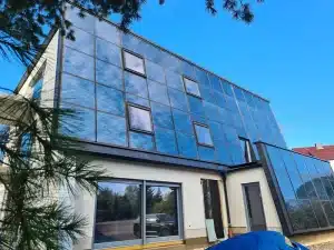 Moderne Solarvilla Schlossblick der FASA AG mit großflächiger, blauer Solar-Glasfassade und integrierten Fenstern, aufgenommen aus der Froschperspektive bei blauem Himmel; im Erdgeschoss große Panoramafenster, im Vordergrund ein abgedeckter Materialhaufen und seitlich Bäume am Bildrand.